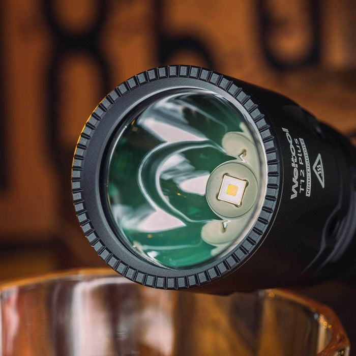 Close-up of the Weltool T12 Plus tactical flashlight showcasing its visible LED bulb and reflective interior, powered by a 21700 battery, aimed at a glass cup.