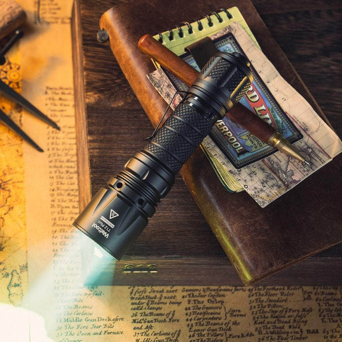 A Weltool T12 Plus tactical flashlight with a 21700 battery and a pen rest on an open notebook atop a leather journal, while a map and compass are in the background.