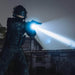 A person in tactical gear stands outdoors at night, aiming a Weltool T12 Plus flashlight powered by a 21700 battery alongside their gun.