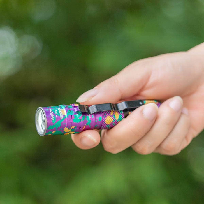 Hand holding a small, colorful Weltool T1Pro V2 Colorful Version LED flashlight with a clip, positioned against a blurred green background.