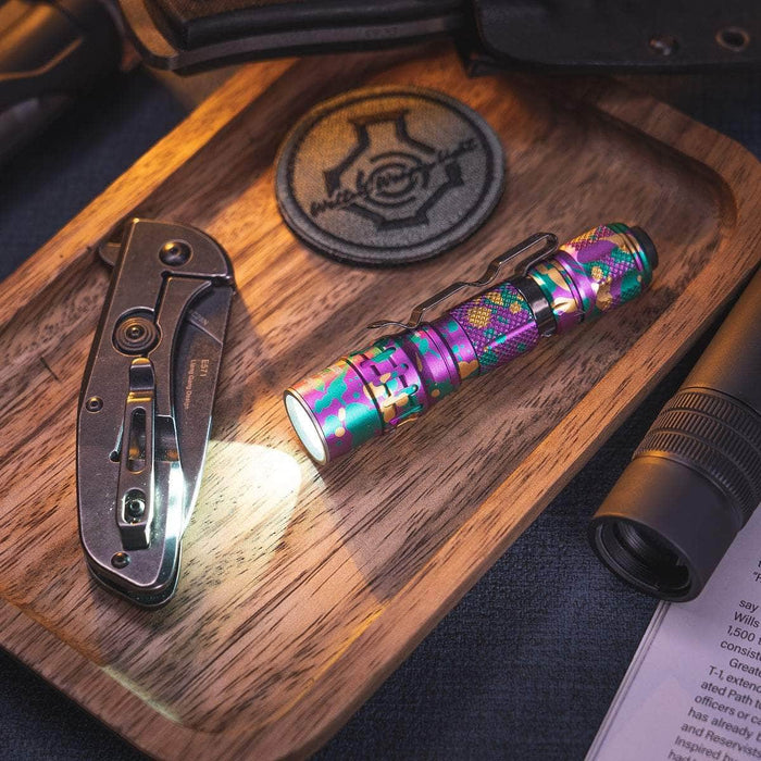 The Weltool T1Pro V2 Colorful Version mini EDC flashlight emits light on a wooden tray next to a folding knife, a decorative patch, and parts of other gadgets.