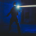 Dressed in tactical gear, a person stands in a dark room holding a baton and the Weltool W3Pro TAC Mini flashlight, its LEP light source cutting through the darkness.